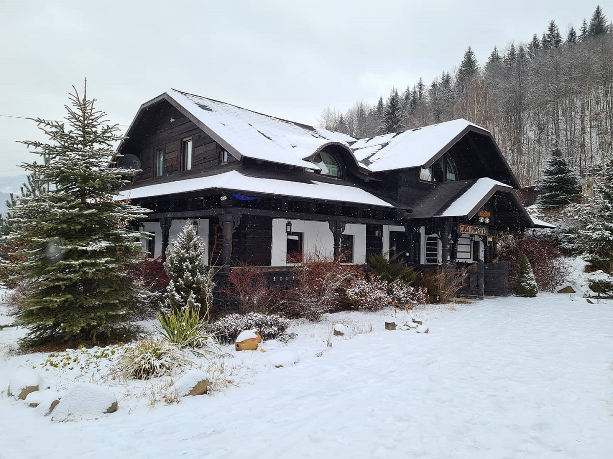 Pensiunea muzeu Casa Poveste, fotografiata din exterior, iarna, imbracata in lemn de culoare ainchisa, cu acoperis nins, si multa zapada in jurul casei - un loc unde să petreci un Crăciun autentic românesc