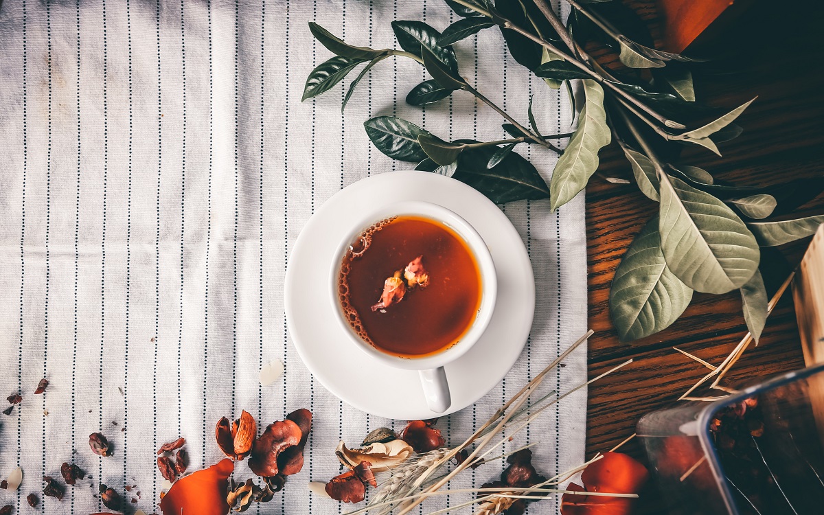 ceasca de ceai fotografiata de sus, asezata pe un stergar alb, alaturi de cracute du frunze verzi si peatle de flori uscate - concept pentru tipuri de ceai