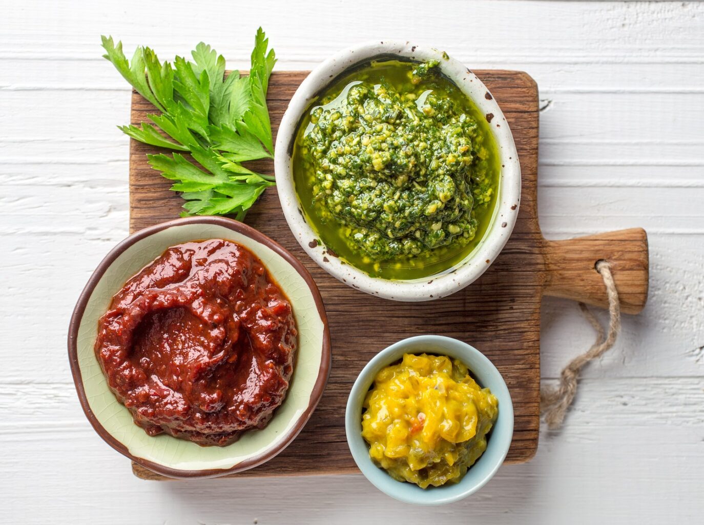 3 boluri cu sosuri diferite, galben, verde și roșu, pe un tocator de lemn, fotografiate de sus. Imagine reprezentativă pentru cele mai populare sosuri din lume