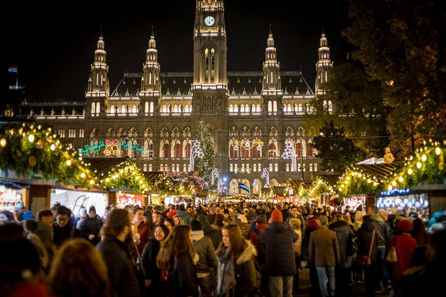 Primaria din Viena fotografiata noaptea, iluminata, iar in plrim plan casute decorate festiv si oameni care se plimbă printre ele la targul de Crăciun la Viena