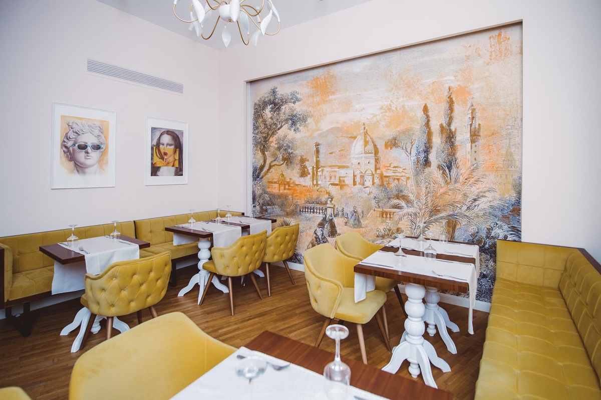 sala de mese de la restaurant lugo, cu mese albe, scaune si ccanapele tapitate galben mustar, un perete pictat si alte 2 tablouri pe alt perete 