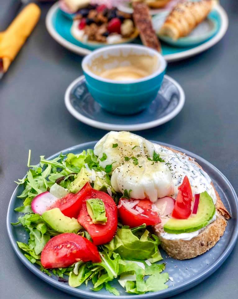 oua posate alaturi de salata verde, rosii si ricotta, pe o farfurie albastra, iar in fundal o ceasca de cafea pentru mic-dejun