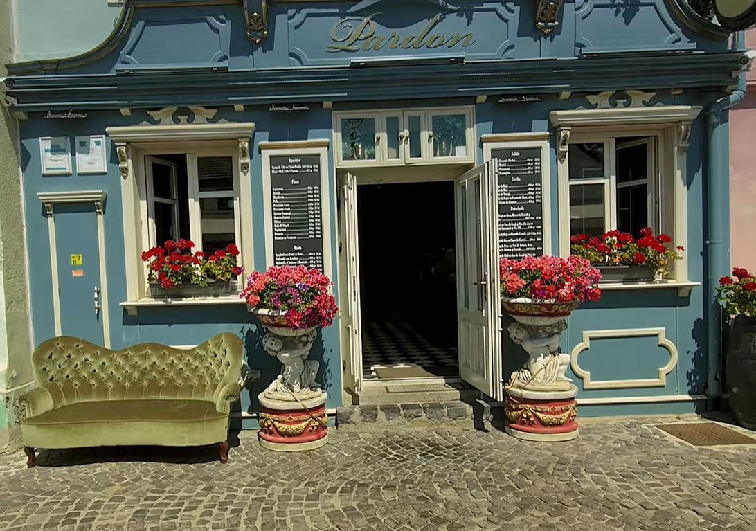 intrarea de la PArdon Cafe Sibiu, cu o canapea tapitata in fata localului, ghivece mari cu flori rosii, pereti albastri si o usa marginata de doua ferestre. Unul dintre cele mai bune restaurante din Sibiu