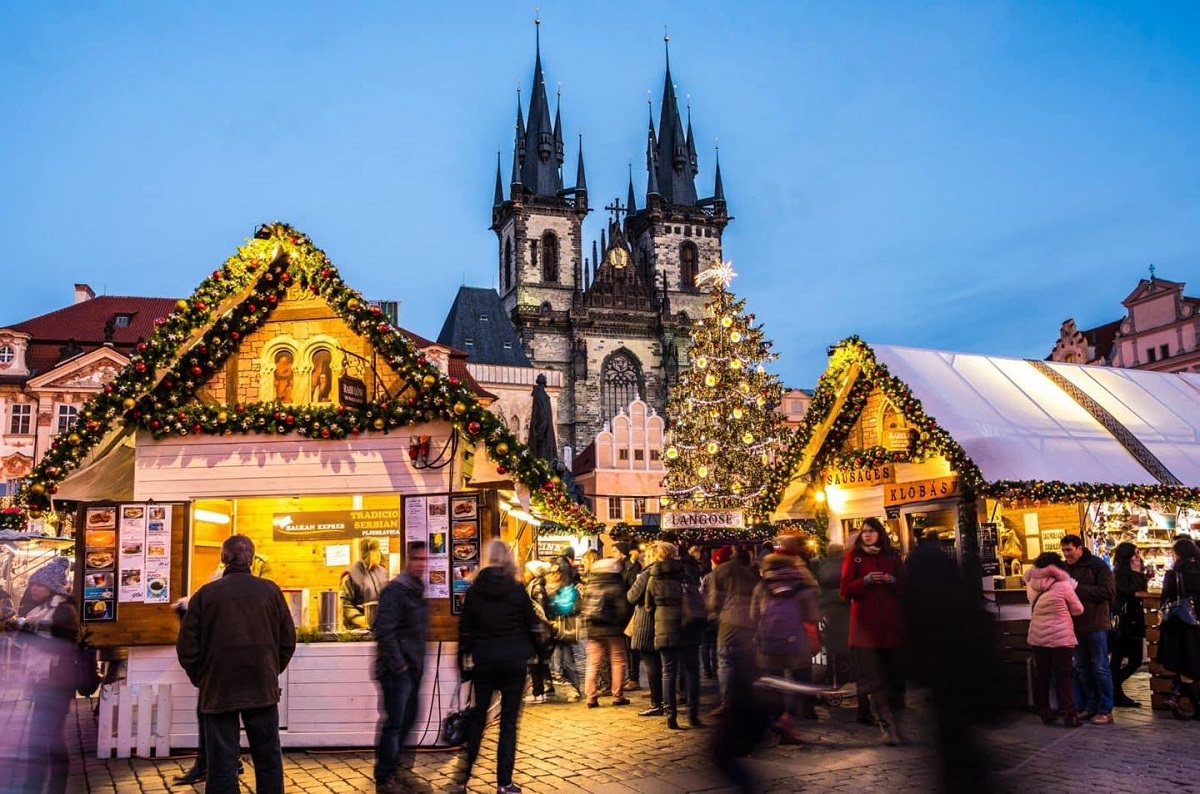 oameni car es eplimbă seara la târgul de Crăciun din Praga, iar in fundal o biserica mare, cu doua turnuri/. Unul dintre cele mai frumoase târguri de Crăciun din Europa.