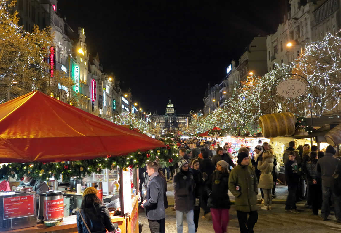 casute care vand suveniruri si cadouri la târgul de Crăciun din Praga, fotografiate noaptea, cu oameni in jur