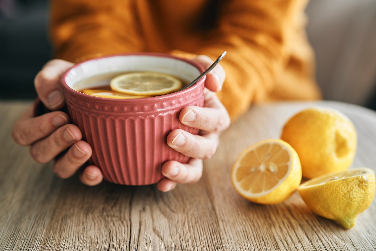close up cu mainile unei femei care tin o ceasca de ceai, roz, cu lamaie in ea si lamai taiate in jumatate langa, pe masa- imagine concept pentru tipuri de ceai si ceainarii frumoase din tara