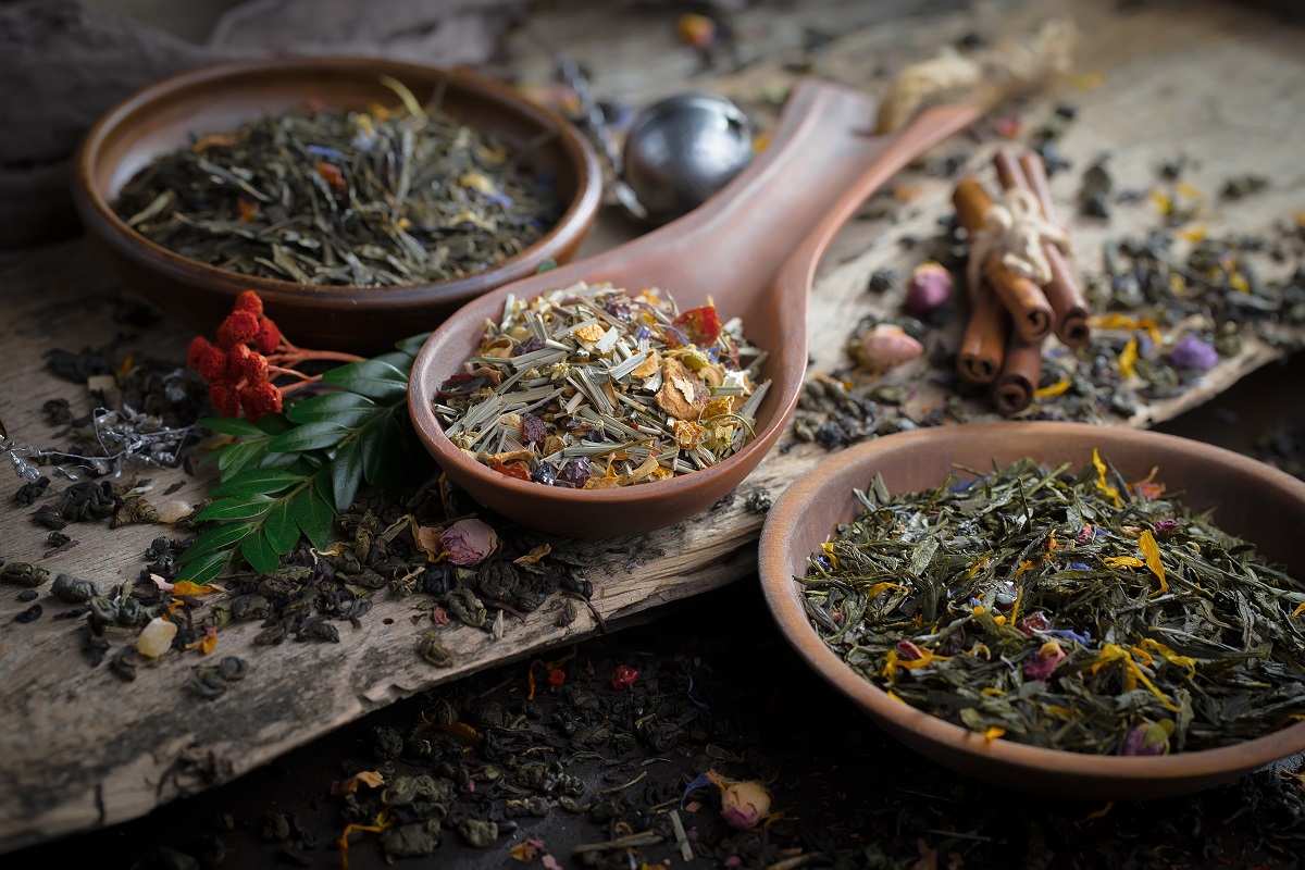 o lingura de lemn alaturi de doua farfurii de lemn, toate pline cu diverse tipuri de ceai uscat din diverse plante, fotografiate in penumbra