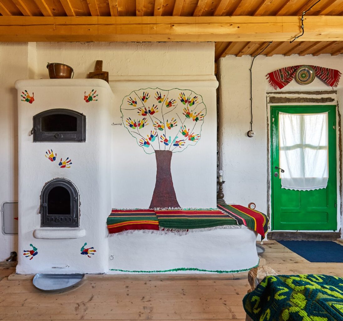 încăpere rustica de la Casa Glod, cu soba din chirpici, alba si pat din chirpici, in prelungirea sobei, cu peretii pictati cu un copac si o usa verde