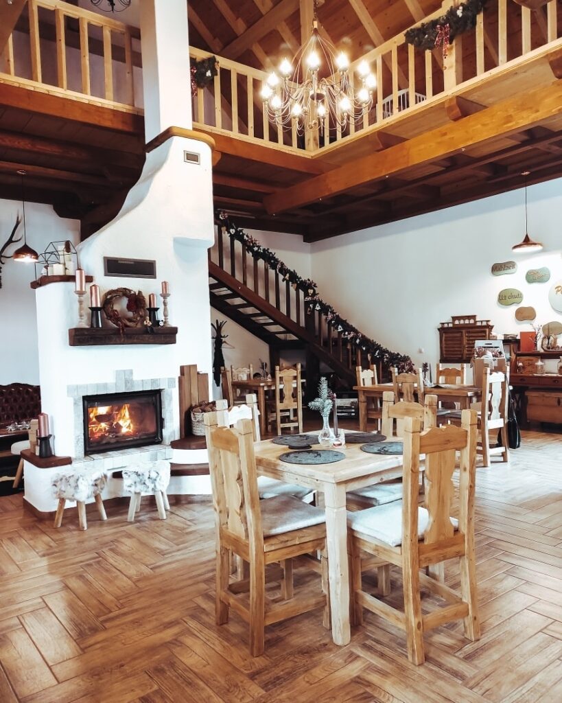 sala de mese de la Casa Trappold, cu semineu alb, rustic, mese si scaune din lemn, si in fundal o scara de lemn care urca la etaj