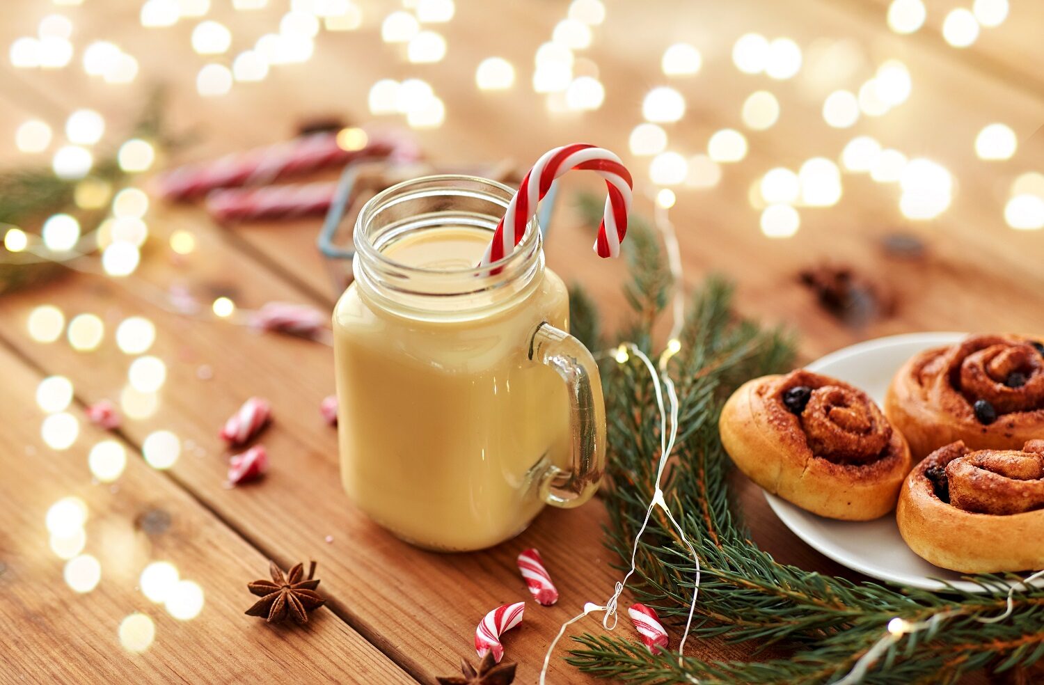 christmas and seasonal drinks concept - eggnog in glass mug with candy cane decoration, cinnamon buns, fir tree brunch and garland lights on wooden background