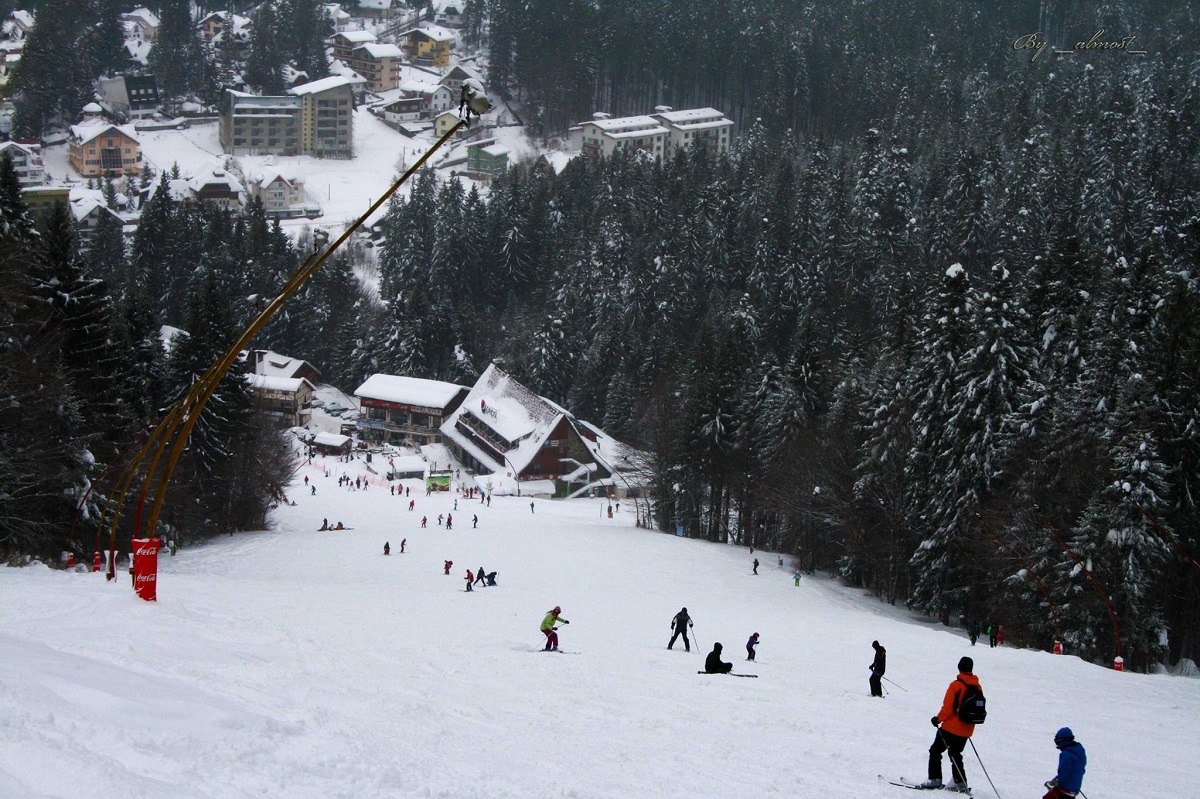 Pârtia de schi Clăbucet din Predeal, fotografiata de sus, cu schiori care coboara la vale, iar in fundal statiunea la poalele muntelui - una din destinații de schi populare din România