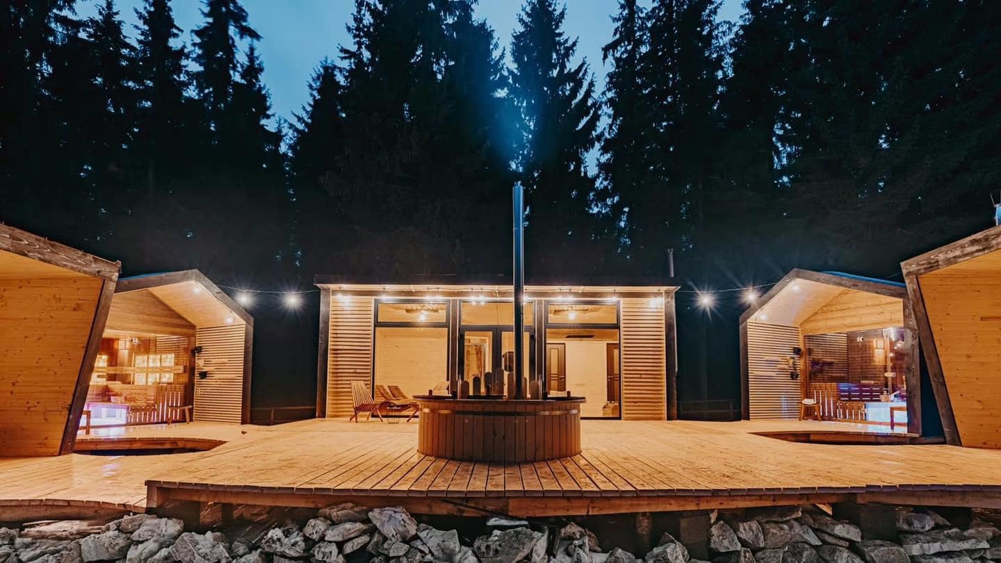 Cabana Bucin & Sauna Village, una din pensiuni cu ciubar, fotografiat seara, in mijlocul unui podet de lemn, inconjurat de 3 cladiri de sauna