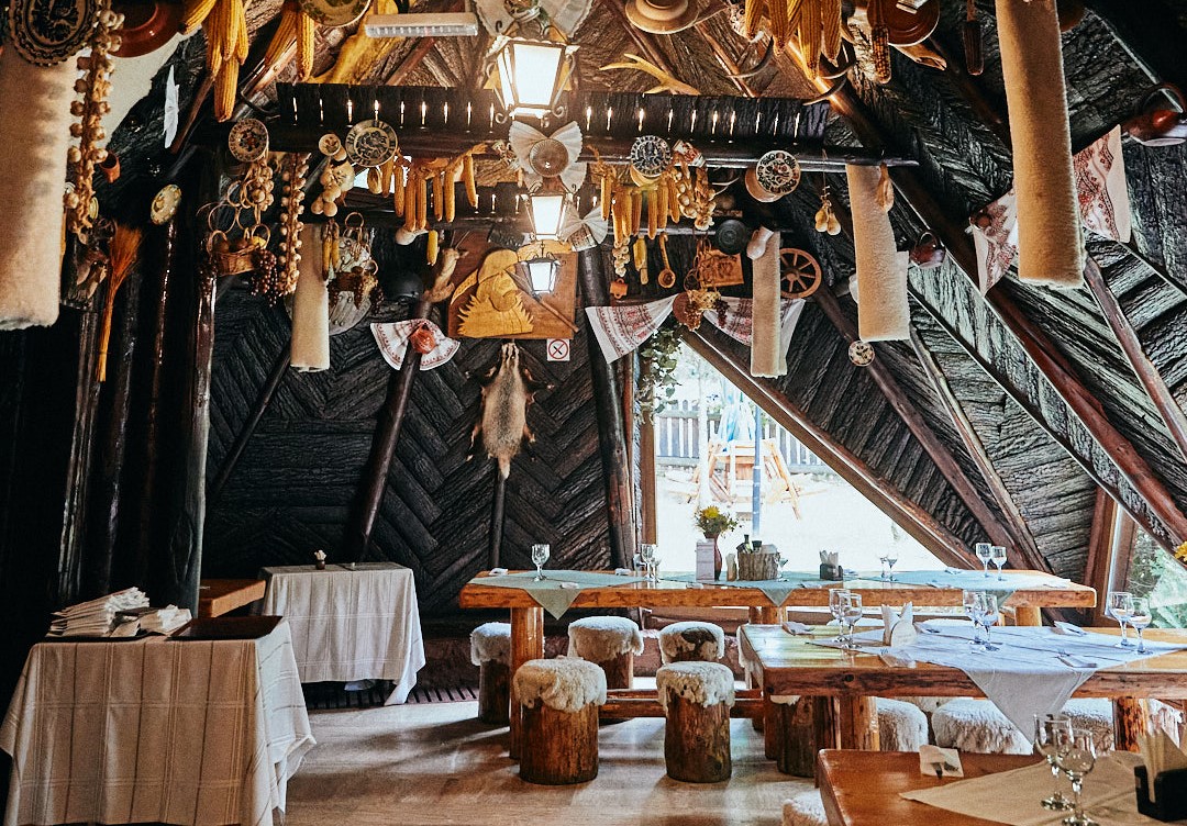 Restaurant rustic Șura Dacilor din Poiana Brasov, cu mese din lemn si tavan inclinat, din lemn, de care stau atarnate carnuri si alimente
