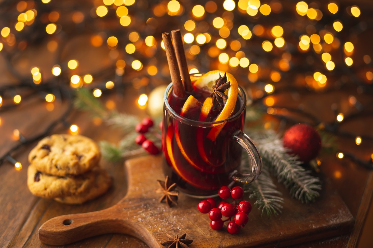 pahar de vin fiert cu portocala si scortisoara, pe o masa de lemn langa doi biscuiti, cu luminite in fundal - cocktailuri de Crăciun