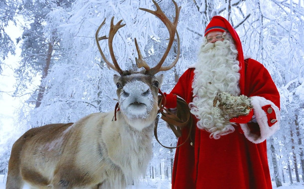 Moș Crăciun alaturi de un Ren iar in fundal copacii acoperiți de zăpadă în Santa Claus Village din Rovaniemi, Laponia