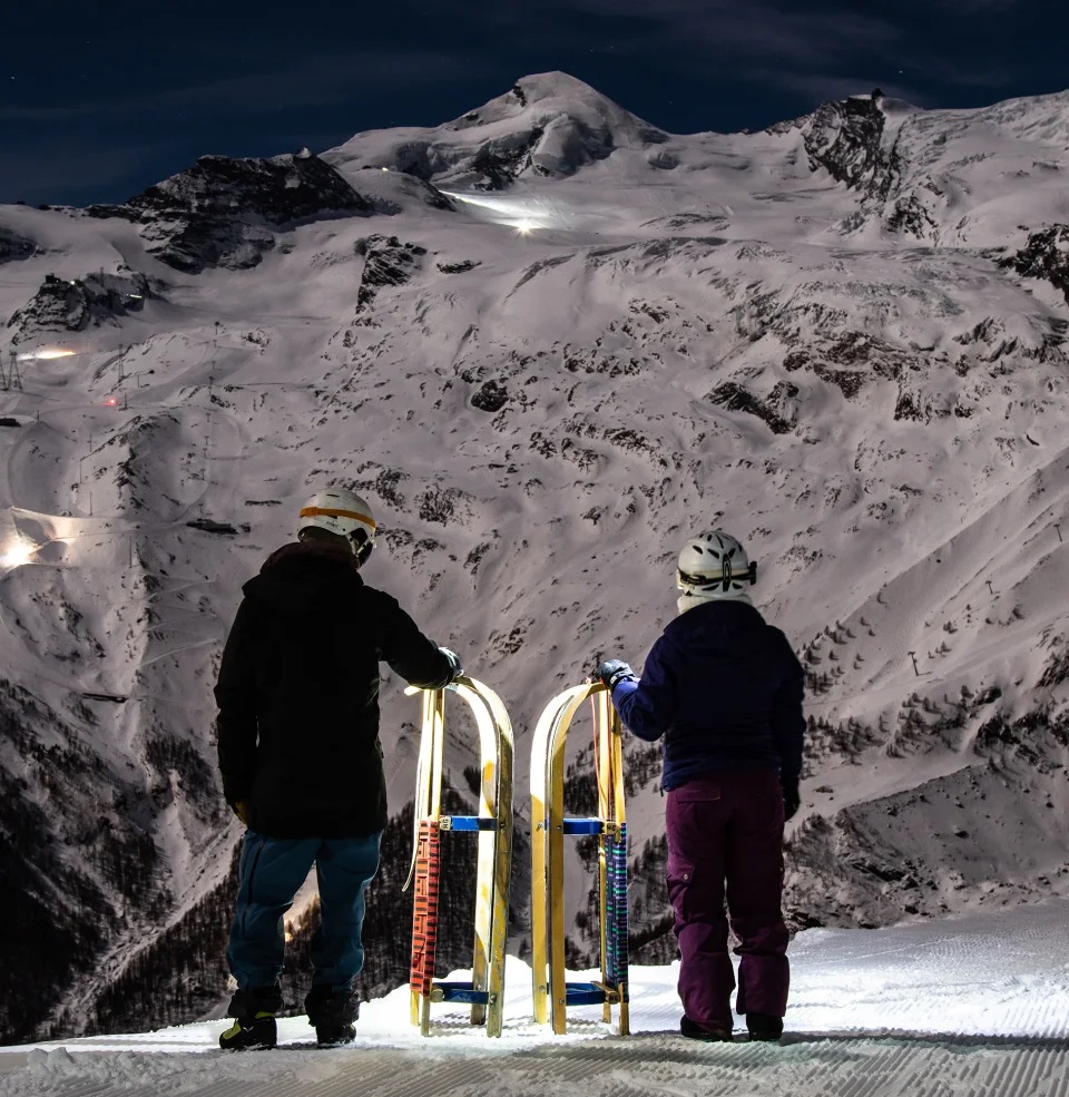 doi oameni cu cate o sanie, care privesc spre muntele acoperit de zapada, fotografiat noaptea, in stațiunea Saas-Fee din Elveția