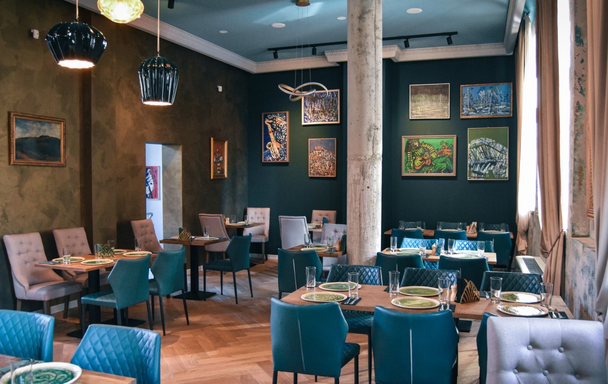 mese in restaurantul Noeme, cu scaune capitonate turcoaz si mese de lemn, de 4 persoane, pereti vopsiti verde inchis, pe care sunt atarnate multe tablouri, si o coloana in mijlocul incaperii 