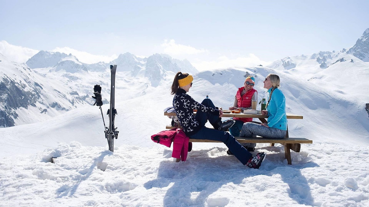 3 femei in costume de schi care iau un picnic la o masa cu bancute din varful muntelui acoperit cu zapada, cu schiurile langa