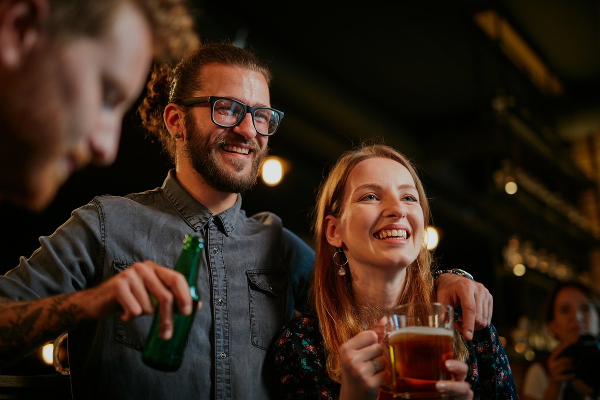 Handsome caucasian man with eyeglasses hugging his girlfriend and talking to her while standing in a pub and drinking beer. Nightlife.