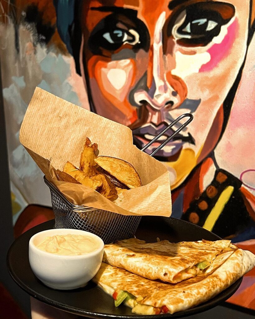 platou cu quesadilla, langa un bol cu sos si altul cu cartofi prajiti, in fata unui tablou cu o negresa, la Tiki Bistro din Iași