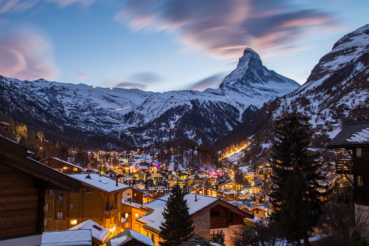 fotografie panoramica asupra statiunii Zermatt, fotografiata seara, cu luminile aprinse la case si in fundal muntii acoperiti de zapada, unde sp mergi la schi în Elveția