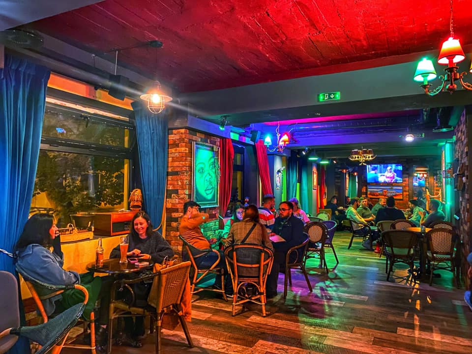 oameni așezați la mese, seara in restaurant Tiki Bistro,  cu geamuri mari si draperii colorate, si tavan din lemn iluminat in mai multe culori, unul din restaurante bune din Iași