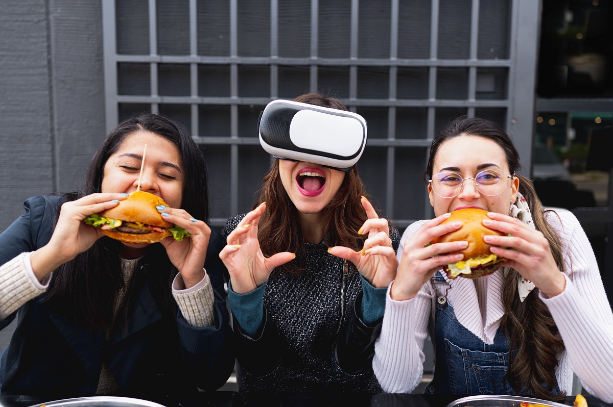 Three young women sitting at table restaurant eating burgers. Woman in the middle wears virtual reality headset simulating to bite and eat.
