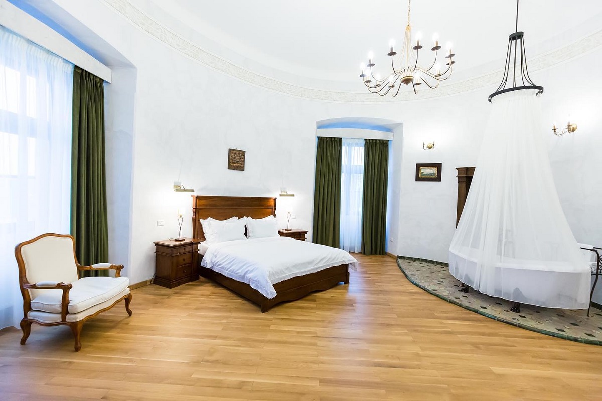 dormitor spatios, cu parchet si pereti albi, ferestre inalte cu draperii, un pat dublu mare, o cada in incapere, acoperita cu un baldachin, si un candelabru elegant, la Castelul Bethlen-Haller, una din destinații romantice din România
