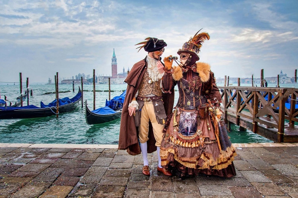 indragostiti costumati si cu masti, fotografiati in fata gondolelor pe apa, la Carnavalul de la Veneția
