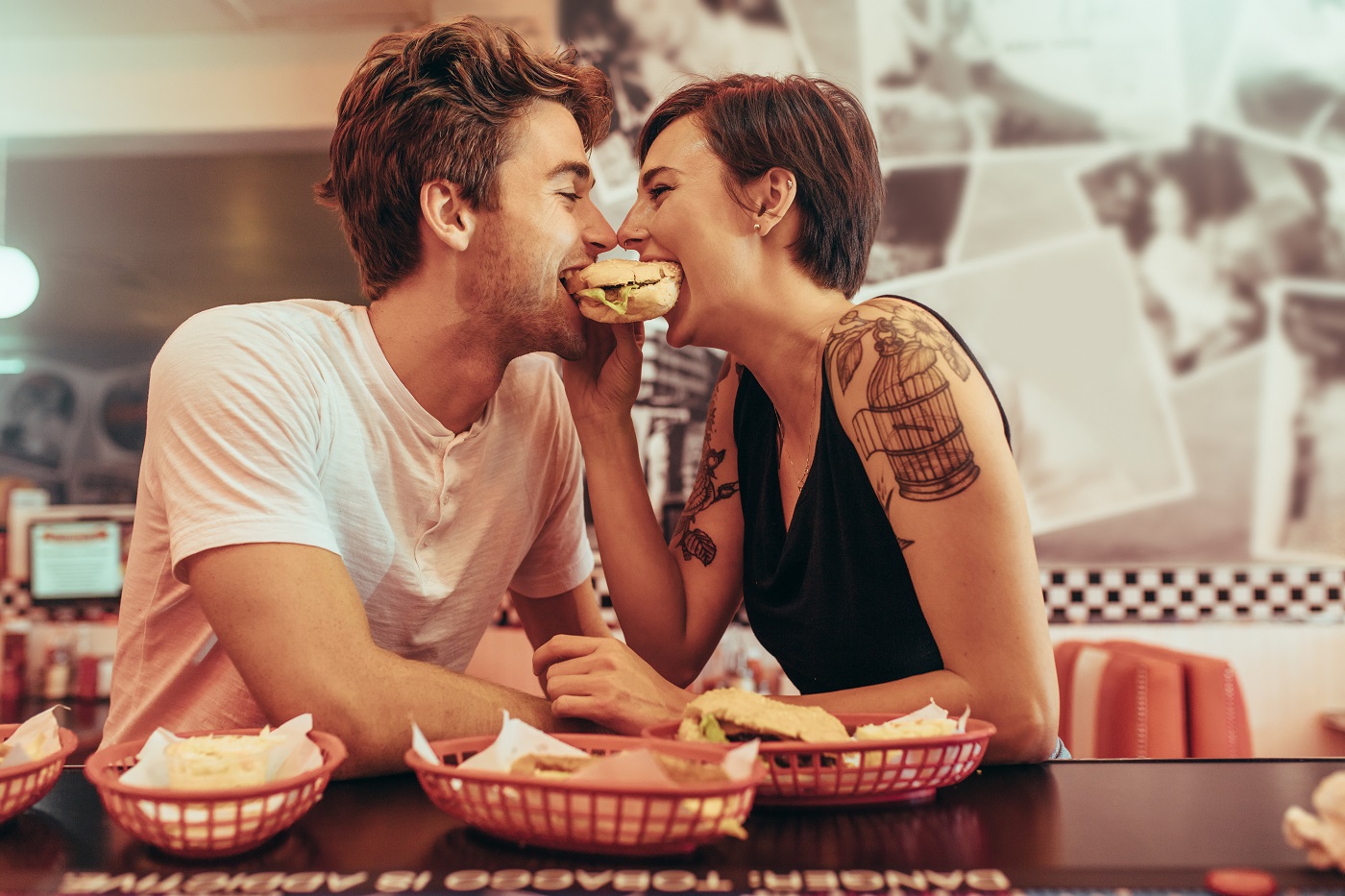 Happy couple at a restaurant eating a burger together looking at each other. Man and woman sitting in a diner with food on the table sharing a burger. - codul bunelor maniere la date