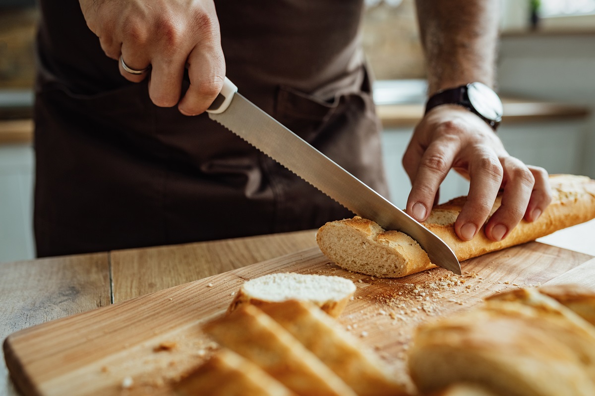close up cu un bucatar care foloseste un cuțit de pâine pentru a felia o bagheta pe un tocator de lemn - unul din tipuri de cuțite