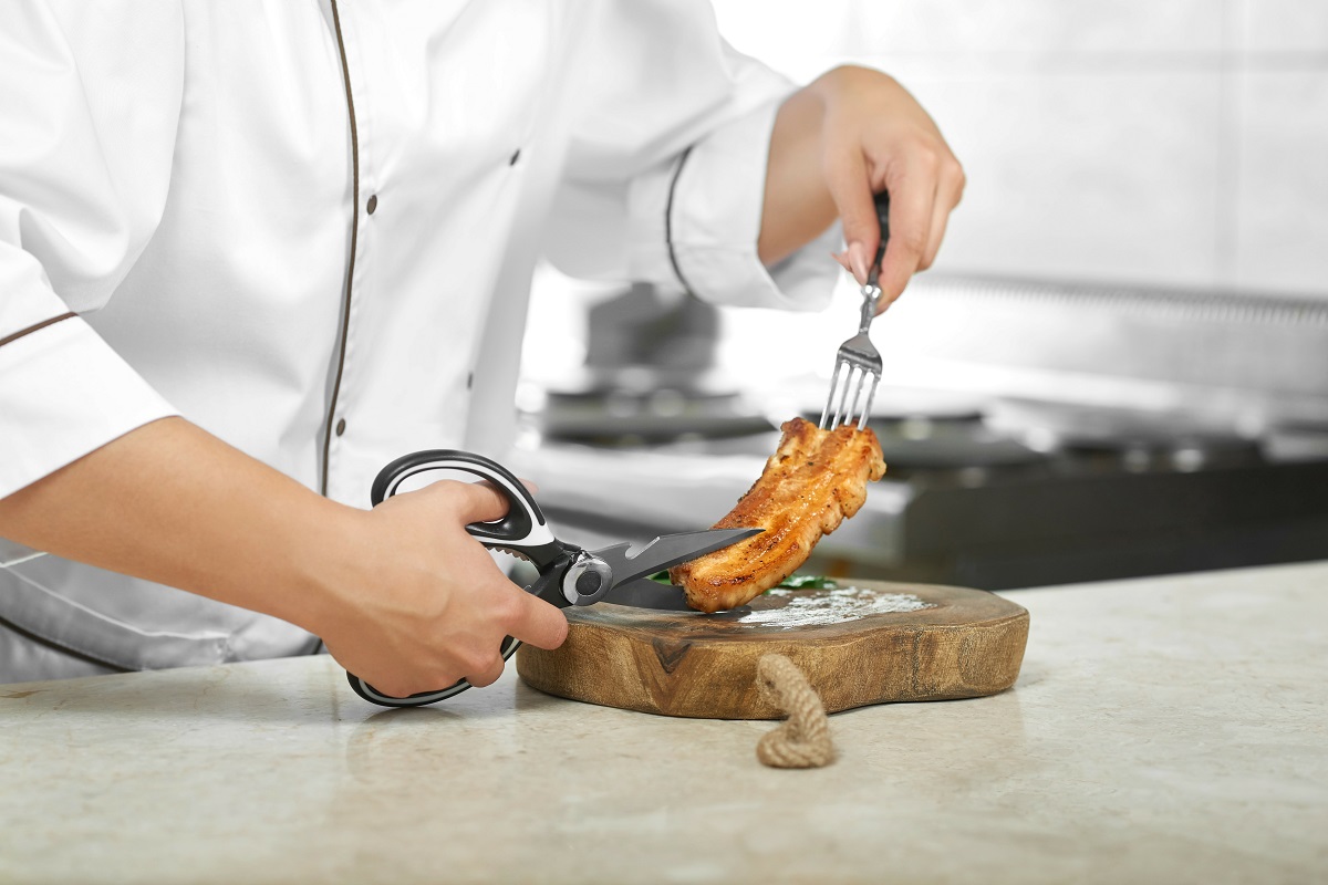 Bucatar care se pregateste sa taie cu foarfeca de bucătărie o felie de pui prăjit, pe un tocător de lemn - unul din tipuri de cuțite de bucătărie