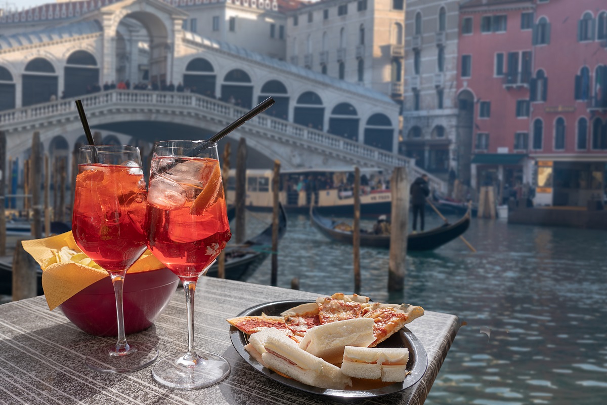 doua pahare de cocktail si o farfurie cu aperitive. iar in fundal podul Rialto peste un canal din Venetia