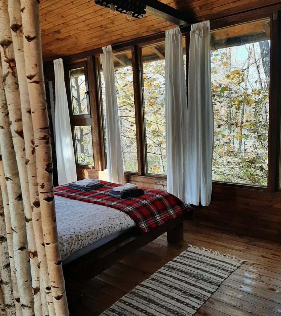 dormitor de la Porumbacu Treehouse, cu un pat asezat in dreptul unor ferestre mari prin care se vede padurea la geam