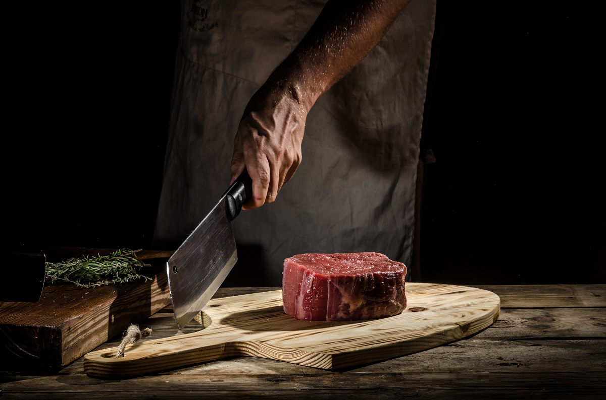 bucata de carne de vita cruda, pe un tocator de lemn, din care un bucatar se pregateste sa taie cu satârul, unul din tipuri de cuțite de bucătărie