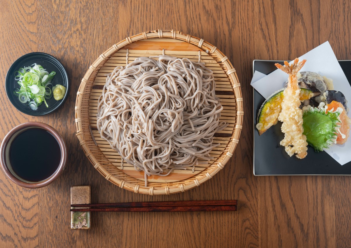 soba noodles asezati pe un platou din bambus, alaturi de 2 boluri mici cu sosuri asiatice si creveti tempura 